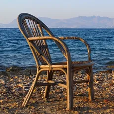 A wicker chair and a car tire on a pebbly beach, with the sea and distant mountains in the background.