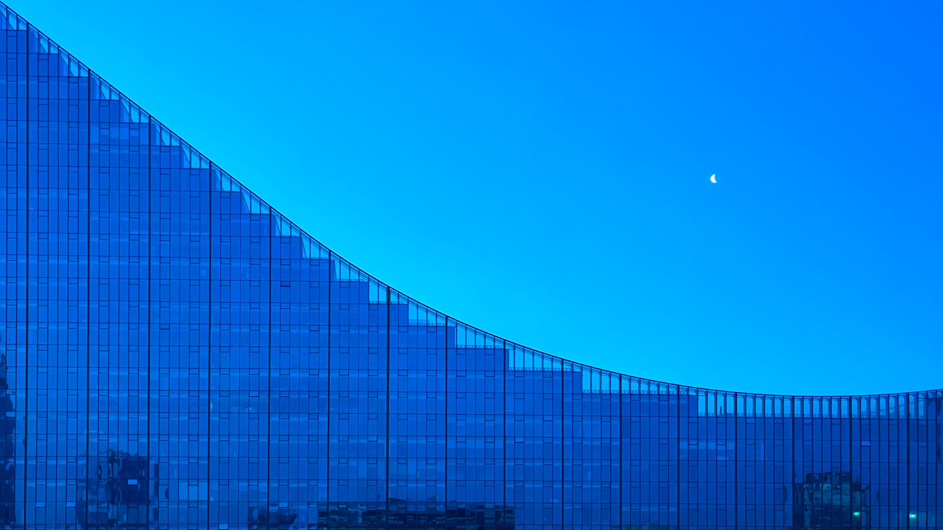 Curved glass building under evening sky with visible crescent moon. Reflections and silhouettes add detail to the modern architectural design.